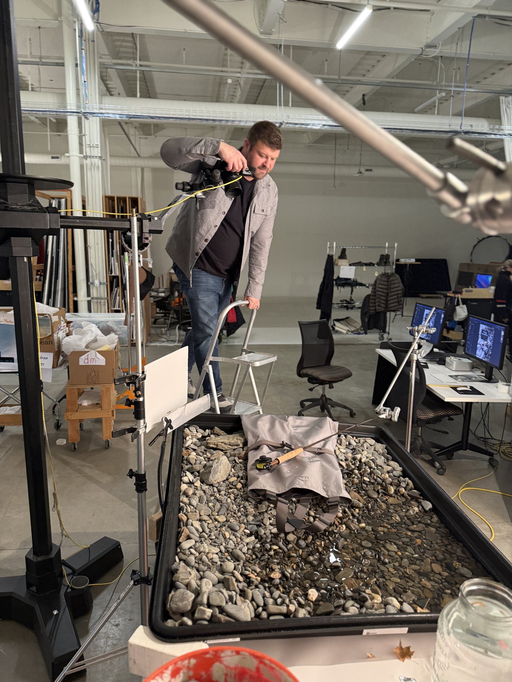 Rory Scranton behind a boom-mounted camera rig, tilted down over a studio product tray of river rocks and water with a fly rod and waders bag staged in it, behind-the-scenes photography.