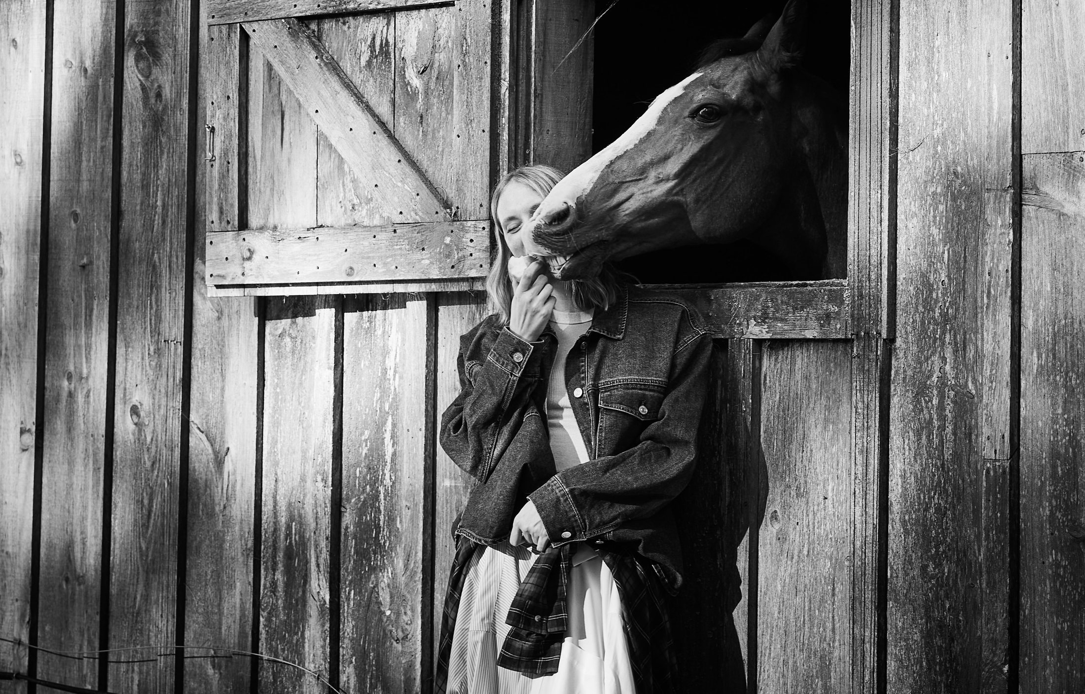 Black and white portrait of a woman laughing with a horse nuzzling her face through a barn stable window.