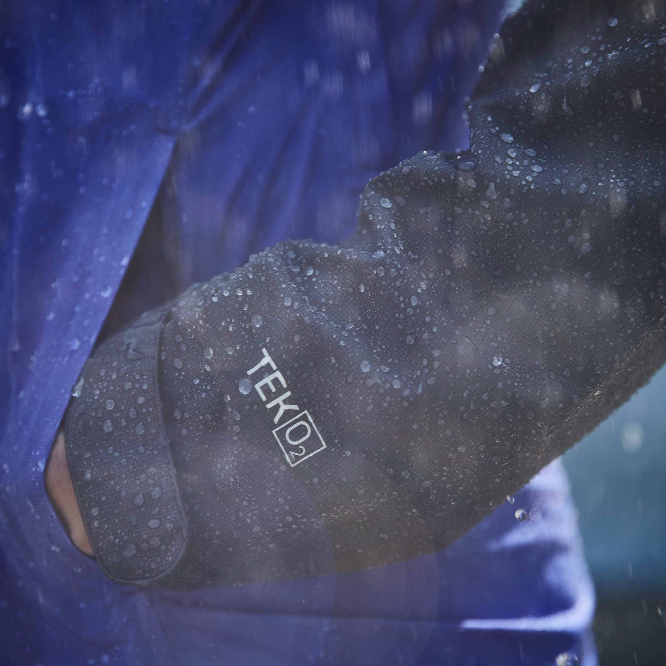 Close-up of a TEK O2 label on a black rain jacket sleeve, rain droplets on fabric.