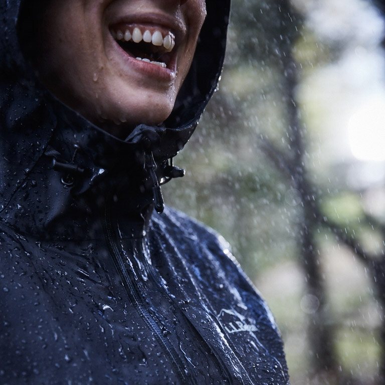 Laughing hiker in a black rain jacket hood, rain falling.