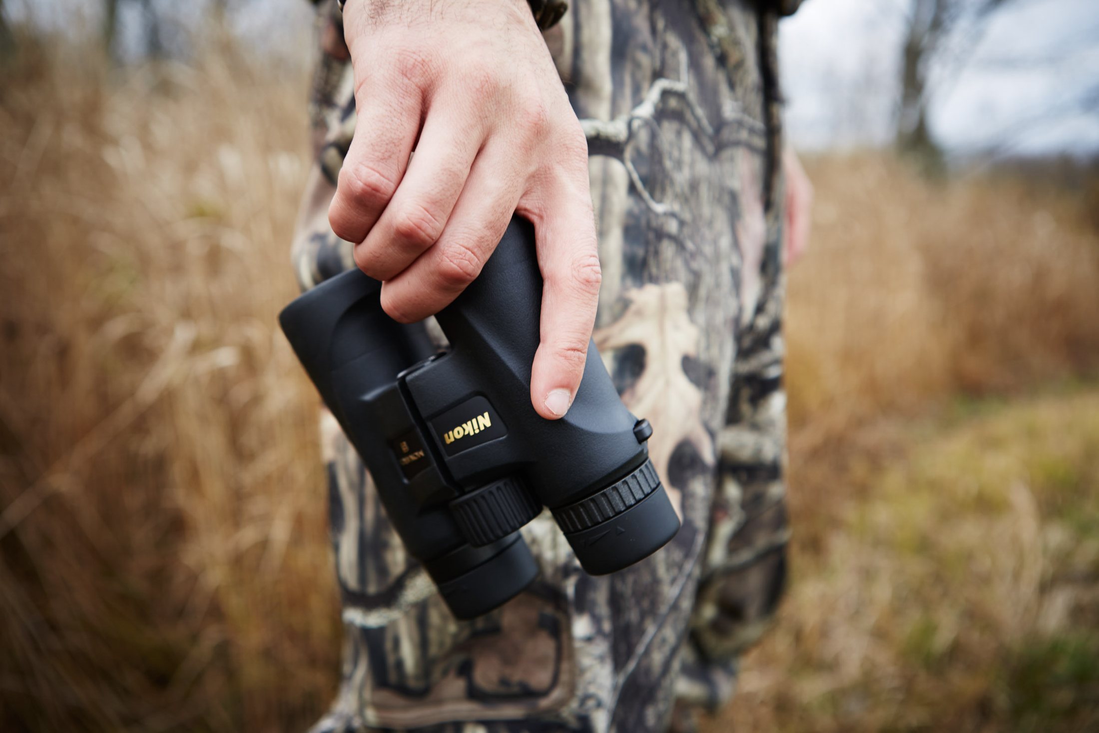 Hand gripping a pair of binoculars, camo pants and autumn field behind, hunting lifestyle photography by Rory Scranton.
