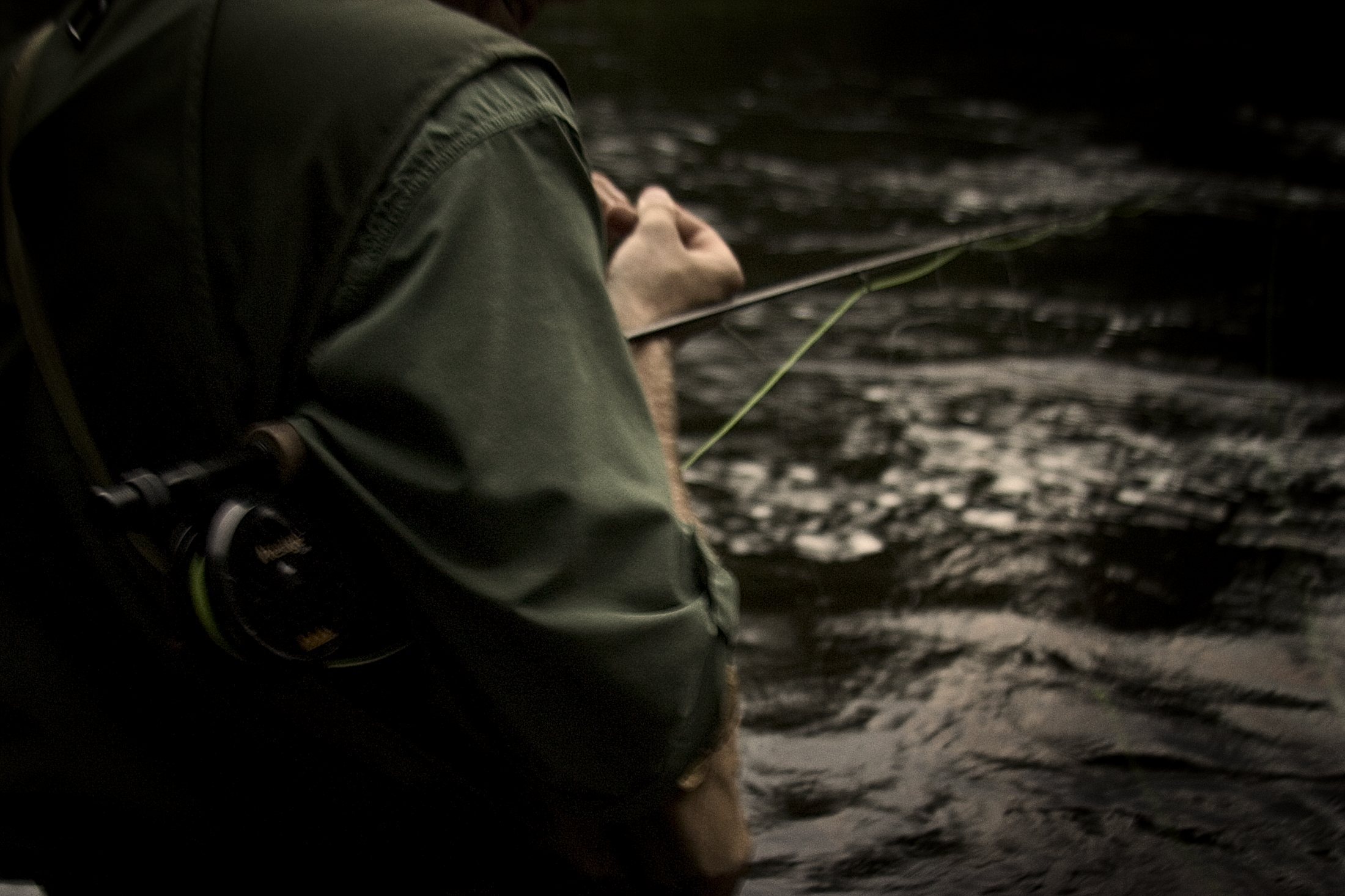 Close, dark detail of an angler's shoulder and fly reel against the river, fly fishing lifestyle photography by Rory Scranton.