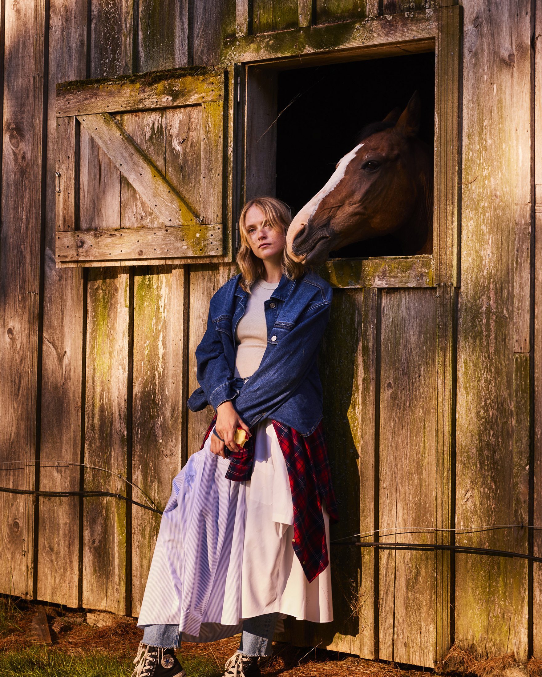 Woman leaning against a weathered barn doorway holding an apple, with a horse reaching its head out through the stable window above her, equestrian lifestyle photography by Rory Scranton for the Norumbega Farm project.