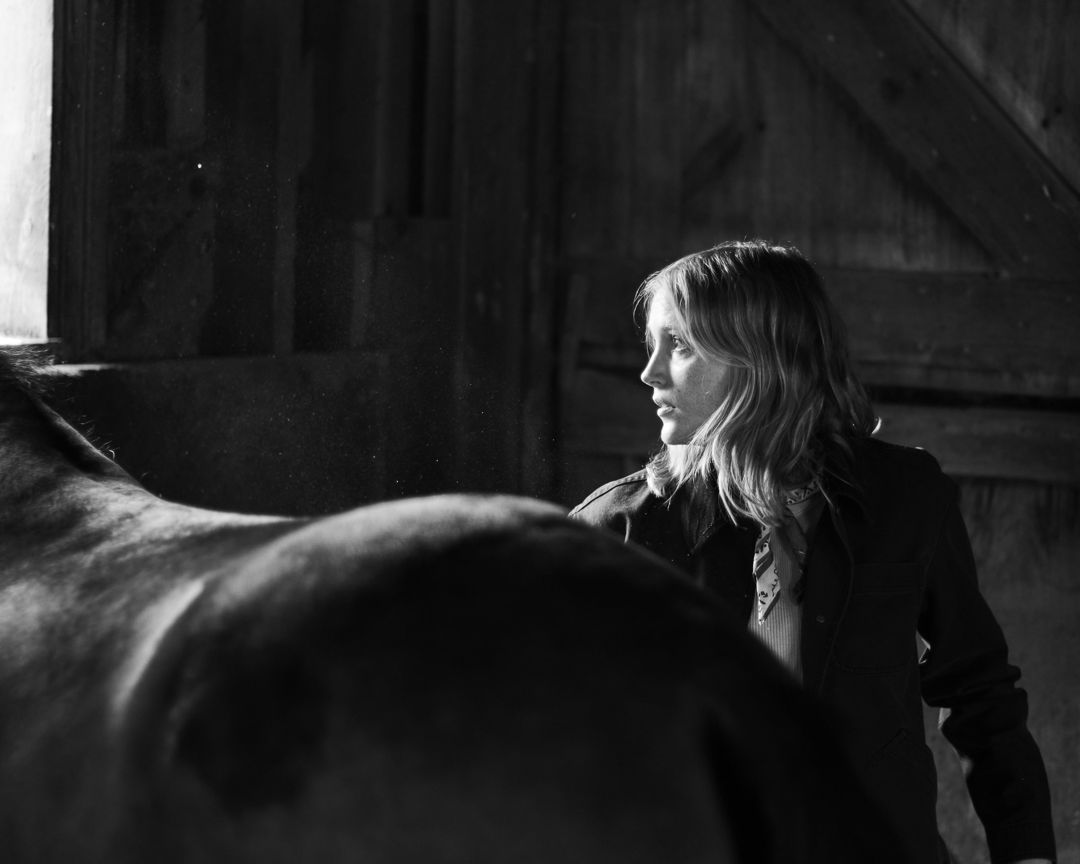 Black and white portrait of a woman standing beside a horse inside a barn, dust visible in a shaft of window light, equestrian lifestyle photography by Rory Scranton for the Norumbega Farm project.