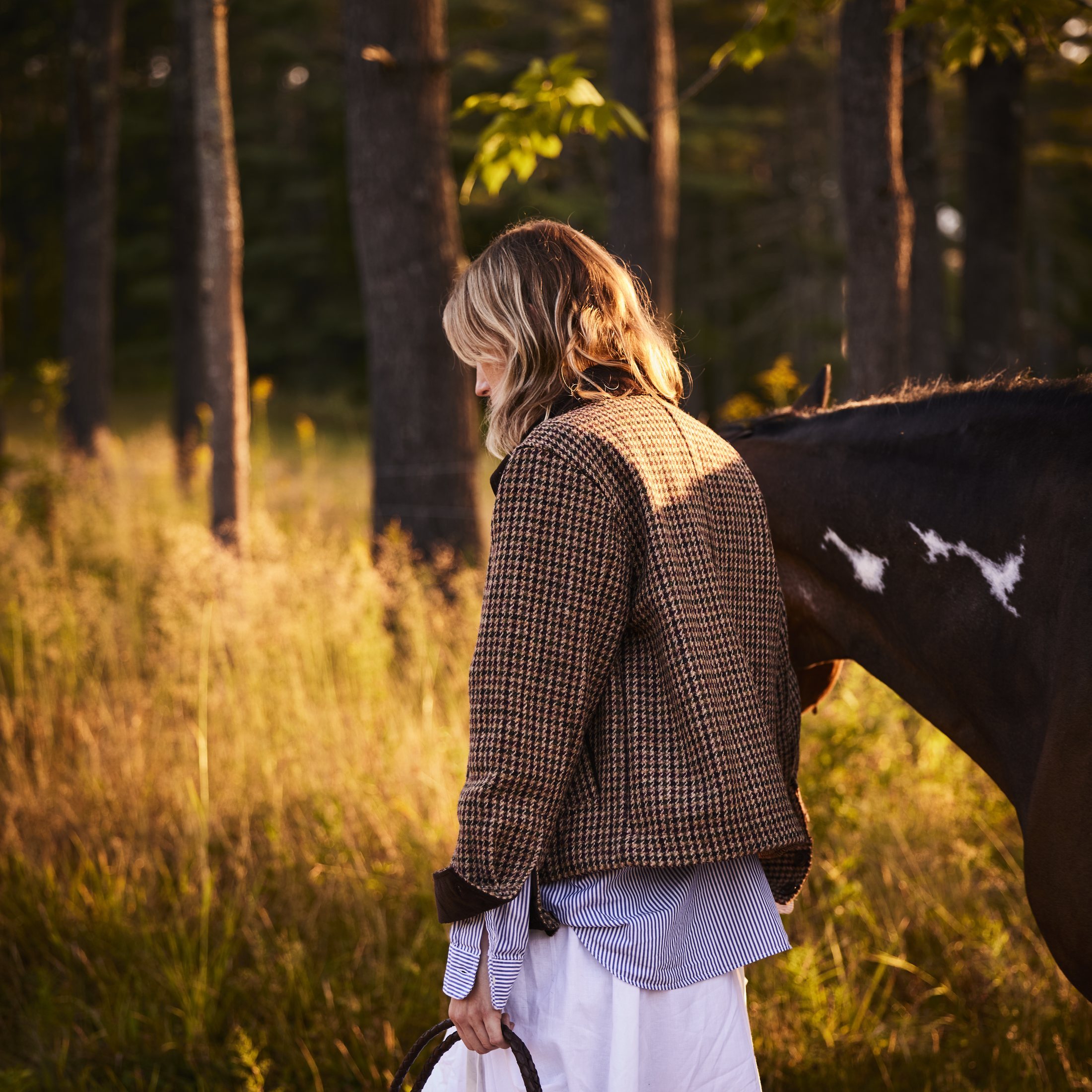 Woman walking beside a horse at golden hour, holding braided leather reins, seen from behind, equestrian lifestyle photography by Rory Scranton for the Norumbega Farm project.