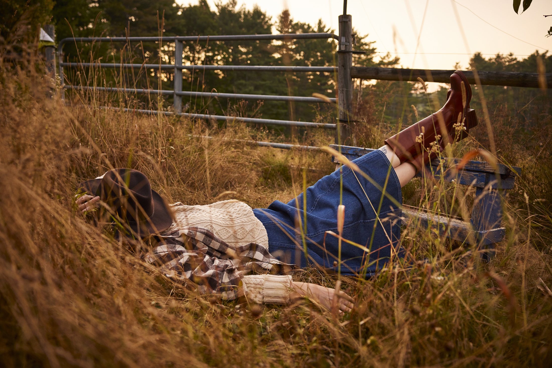 Woman resting in tall golden grass with her hat over her face, boots propped on a weathered fence rail, equestrian lifestyle photography by Rory Scranton for the Norumbega Farm project.